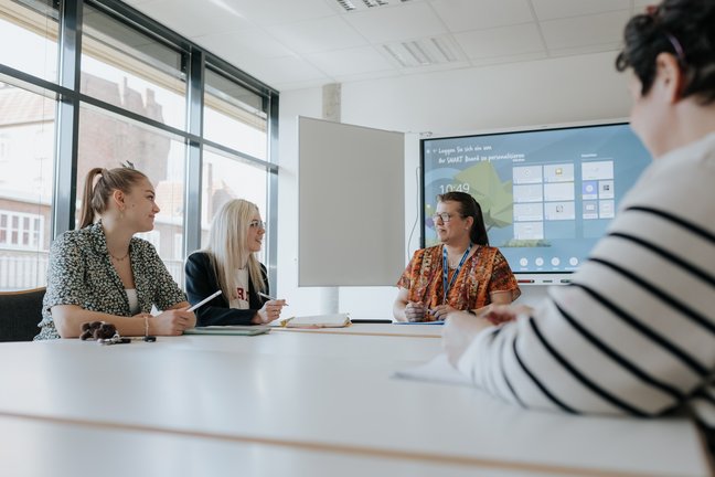 Vier Personen sitzen in einem Besprechungsraum um einen Tisch herum und diskutieren vor einem Präsentationsbildschirm im Hintergrund.