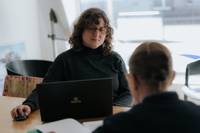 Zwei Personen sitzen an einem Tisch; eine benutzt einen Laptop, während die andere in einem hellen Raum Dokumente überprüft.