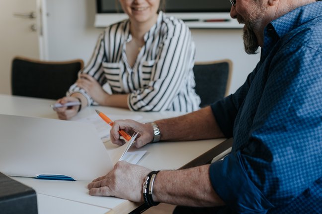 Zwei Personen sitzen an einem Tisch und besprechen Dokumente; einer schreibt mit einem orangefarbenen Stift, beide lächeln.