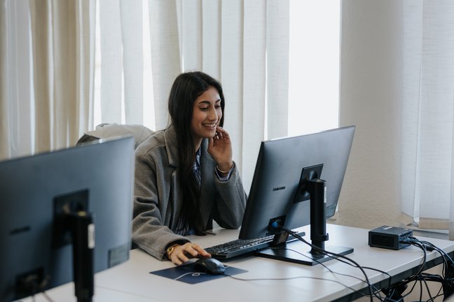Eine Frau lächelt, während sie in einem hellen, modernen Büro an einem Computer arbeitet.