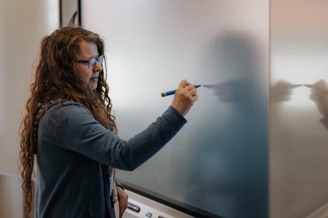 Eine Frau mit langen Haaren schreibt in einem Klassenzimmer mit einem blauen Marker auf eine Tafel.
