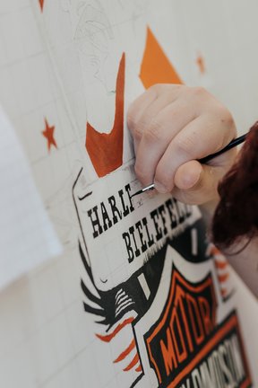 Eine Person malt mit einem kleinen Pinsel das Wort HARLEY auf ein detailliertes Harley-Davidson-Schild.