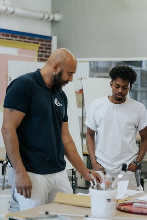 Zwei Männer stehen an einem Tisch mit Malutensilien und diskutieren in einer hellen, kreativen Atelierumgebung über etwas.