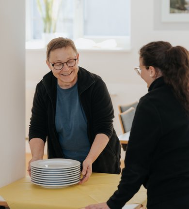 Zwei Personen lächeln, während sie einen Stapel Teller auf einen mit einem gelben Tischtuch bedeckten Tisch stellen.