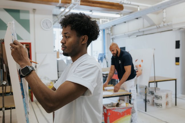 Zwei Künstler arbeiten in einem hellen Atelier; einer malt an einer Staffelei, der andere arbeitet an einem Tisch im Hintergrund.