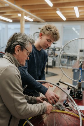 Ein älterer Mann und ein junger Mann reparieren gemeinsam ein Fahrradrad in einer Werkstatt.