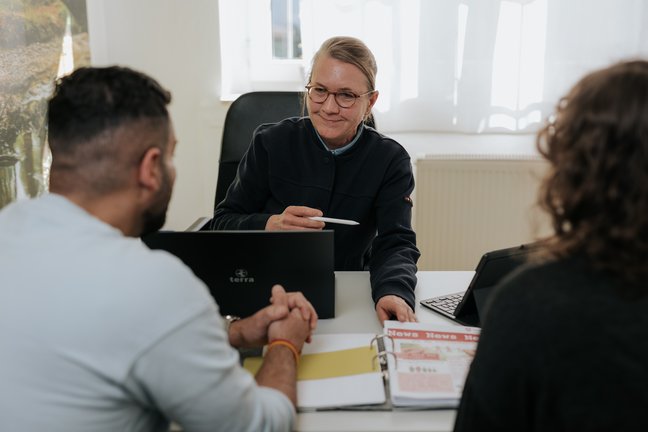 Eine Frau mit Brille unterhält sich mit zwei Personen an einem Schreibtisch mit einem Laptop und einem aufgeschlagenen Aktenordner.