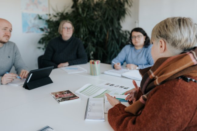 Vier Personen sitzen um einen Tisch mit Notizbüchern und Papieren und diskutieren in einem hellen Raum.