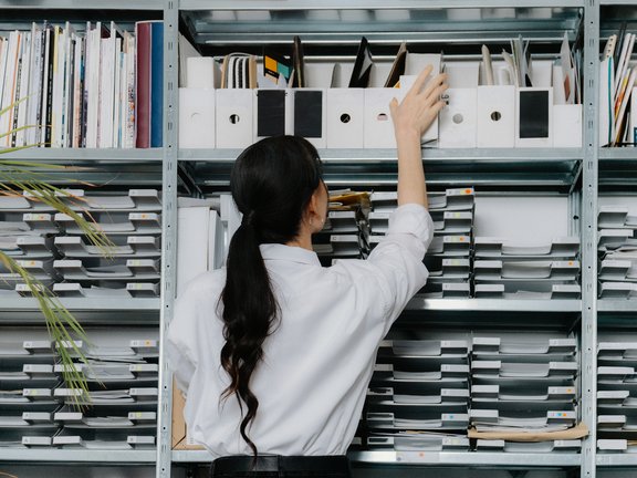 Eine Frau in einem weißen Hemd greift nach einem Ordner in einem Regal, das mit geordneten Dokumenten und Ordnern gefüllt ist.