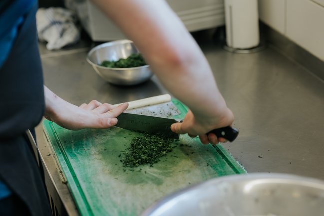Eine Person schneidet frische Kräuter auf einem grünen Schneidebrett in einer Küche, in der Nähe stehen Metallschalen.