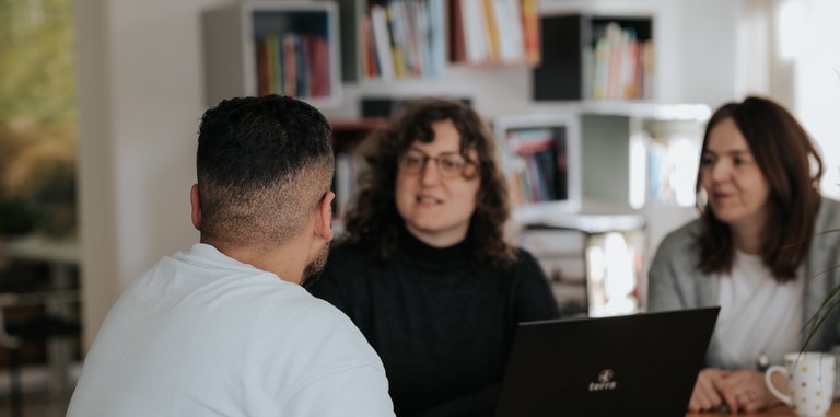 Drei Personen sitzen an einem Tisch und unterhalten sich in einem hellen Raum mit einem Laptop, Papieren und Bechern.