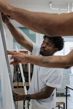 Ein Mann in einem weißen Hemd arbeitet an einer Leinwand, während eine andere Person mit dem Arm auf das Kunstwerk zeigt.