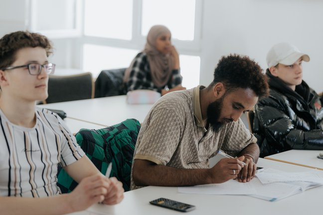 Vier Schüler sitzen an den Tischen in einem Klassenzimmer, ein Schüler schreibt in ein Heft.