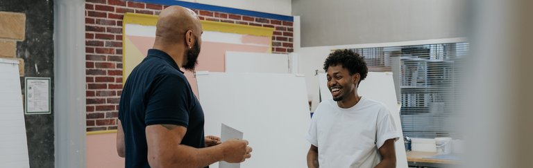 Zwei Männer stehen und lächeln, während sie in einem hell erleuchteten Atelier an Kunstprojekten arbeiten.