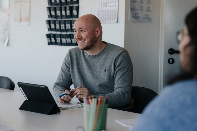 Ein lächelnder Mann in einem grauen Pullover sitzt während einer Besprechung an einem Schreibtisch mit einem Tablet und einem Stift.