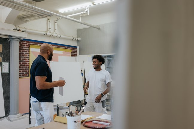 Zwei Männer in einem Kunstatelier lachen und unterhalten sich, während sie neben einer leeren Leinwand und Malutensilien stehen.