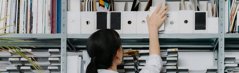 Eine Frau in einem weißen Hemd greift nach einem Ordner in einem Regal, das mit geordneten Akten und Dokumenten gefüllt ist.
