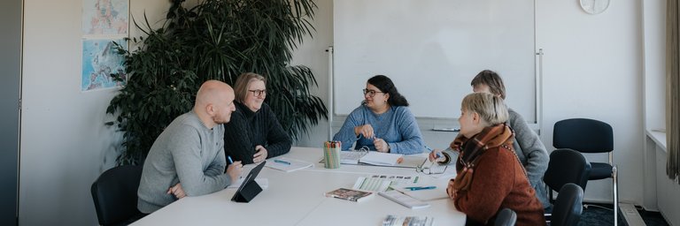 Fünf Personen sitzen in einem Besprechungsraum um einen Tisch herum, unterhalten sich und schauen auf Dokumente und einen Laptop.