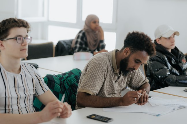 Schüler sitzen an den Tischen in einem Klassenzimmer; einer schreibt, während die anderen aufmerksam zuhören.