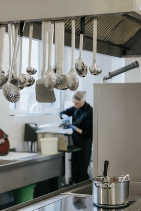Metallkellen hängen über einer Herdplatte mit einem Topf; im Hintergrund arbeitet eine Person in einer Großküche.