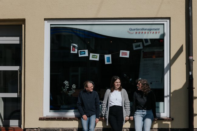 Drei Frauen sitzen und unterhalten sich vor einem großen Fenster, in dem Karten und Schilder ausgestellt sind.