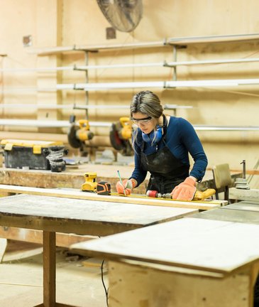Eine Frau misst mit Handschuhen und Schutzausrüstung Holz an einer Werkbank in einer Schreinerei aus.