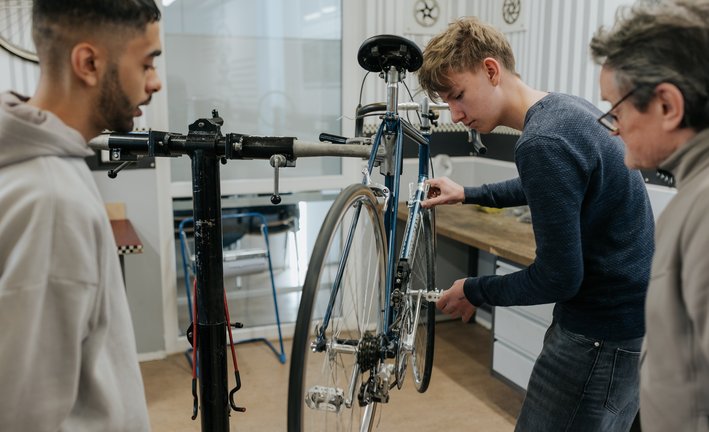 Drei Personen reparieren gemeinsam ein Fahrrad in einer Werkstatt, wobei sie sich auf die Einstellung der Pedale und Räder konzentrieren.