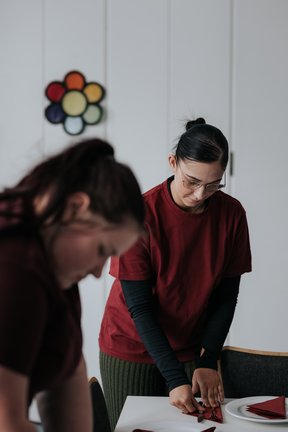 Zwei Frauen falten Servietten an einem Tisch, beide tragen rote Hemden, in einem hellen Raum mit weißen Wänden.