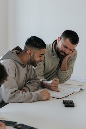 Zwei Männer, die lächelnd an einem Tisch sitzen und mit einem Notizbuch und einem Taschenrechner arbeiten.