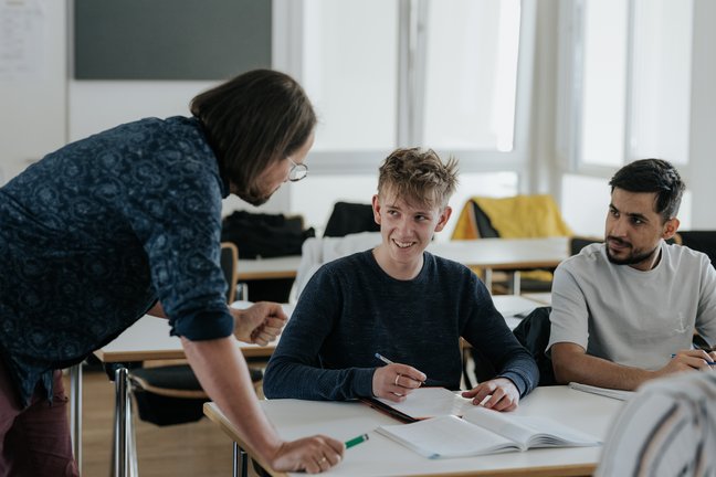 Lehrer im Gespräch mit zwei Schülern an einem Schreibtisch; ein Schüler lächelt und hält einen Stift, der andere hört zu.