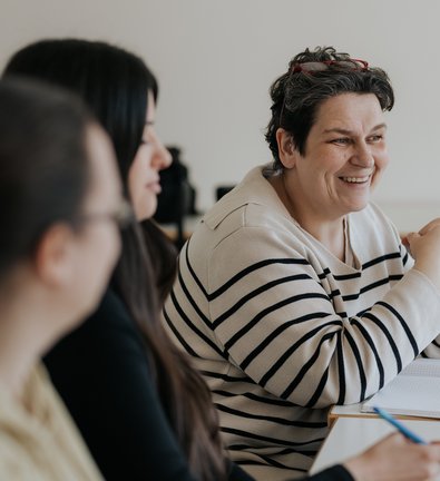 Drei Frauen sitzen an einem Tisch mit Notizbüchern, eine lächelt und ist in ein Gespräch vertieft.