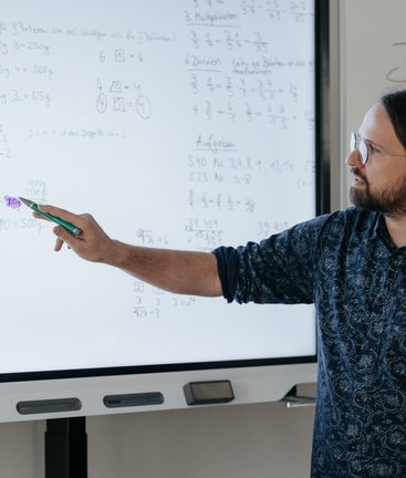 Ein Mann zeigt auf ein digitales Whiteboard, auf dem handschriftliche mathematische Notizen und Gleichungen angezeigt werden.