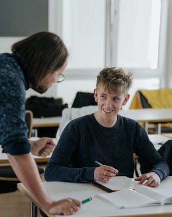 Ein Lehrer spricht mit zwei männlichen Schülern, die in einem hellen Klassenzimmer an einem Schreibtisch sitzen und in Hefte schreiben.