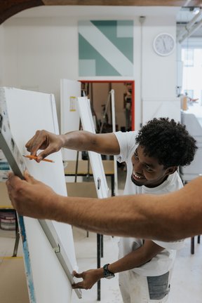 Eine lächelnde Person in einem weißen Hemd misst eine Leinwand in einem Kunstatelier mit Staffeleien und einer Uhr an der Wand aus.
