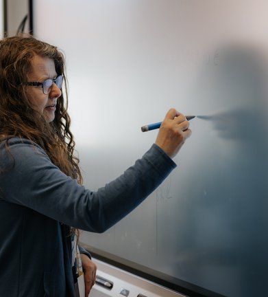 Eine Person mit langen Haaren und Brille schreibt mit einem blauen Marker auf eine große Tafel.