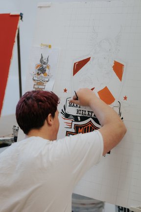 Eine Person mit kurzen Haaren zeichnet ein von Harley-Davidson inspiriertes Design auf ein großes Rasterpapier.