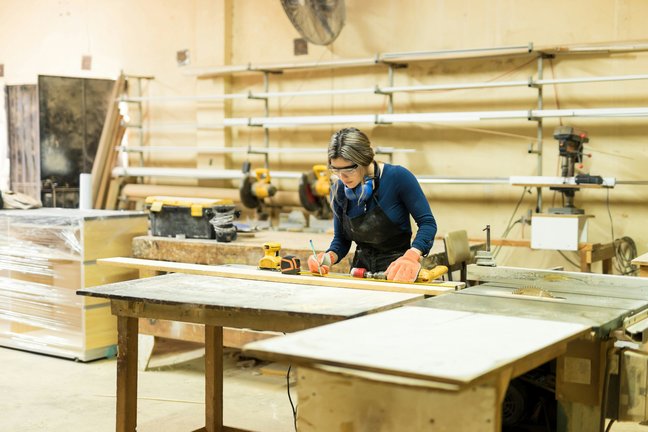 Eine Frau in einer Werkstatt misst und markiert Holz auf einem Tisch, umgeben von Schreinereiwerkzeugen und -geräten.