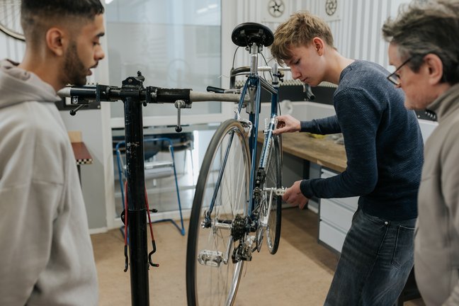 Drei Personen stehen um ein Fahrrad auf einem Reparaturständer, während eine Person die Pedale des Fahrrads in einer Werkstatt einstellt.