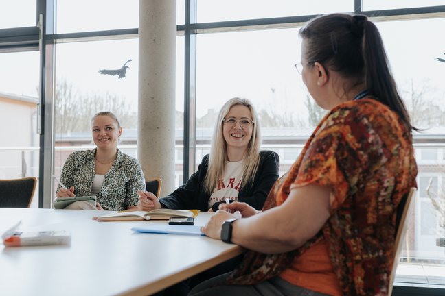 Drei Frauen sitzen lächelnd um einen Tisch und unterhalten sich in einem hellen Raum mit großen Fenstern im Hintergrund.