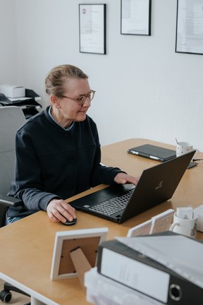 Eine Frau mit Brille arbeitet in einem modernen Büro an einem Schreibtisch mit einem Laptop und lächelt leicht.
