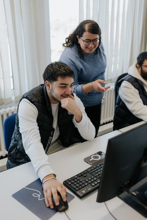 Eine lächelnde Frau hilft einem Mann, der am Schreibtisch einen Computer benutzt, beide wirken glücklich und engagiert.