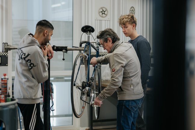 Drei Personen arbeiten gemeinsam an der Reparatur eines Fahrrads in einer Werkstatt mit sichtbaren Werkzeugen und Geräten.