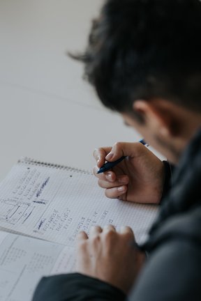 Eine Person schreibt mit einem Stift mathematische Gleichungen in ein Notizbuch, von der Schulter aus gesehen.