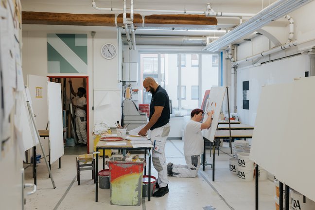 Zwei Personen malen an Staffeleien in einem hellen, modernen Kunstatelier mit Tischen, Material und großen Fenstern.