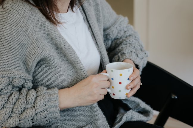 Person in einem grauen Pullover, die eine weiße Tasse mit bunten Punkten hält und auf einem schwarzen Stuhl sitzt.