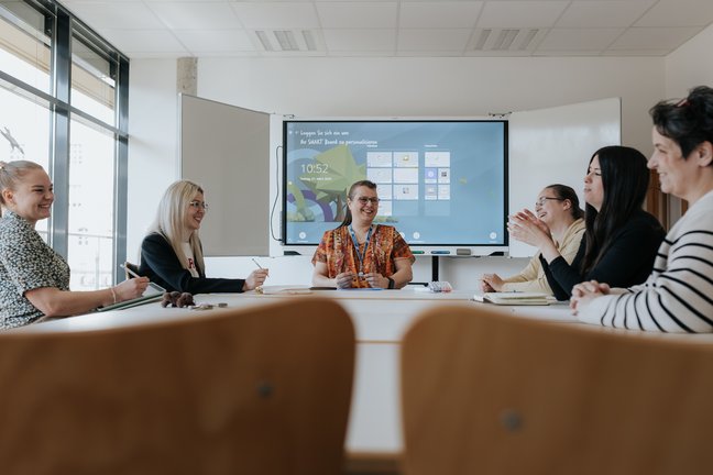 Sechs Frauen sitzen in einem Besprechungsraum um einen Tisch herum, lächeln und sprechen vor einem Bildschirm.
