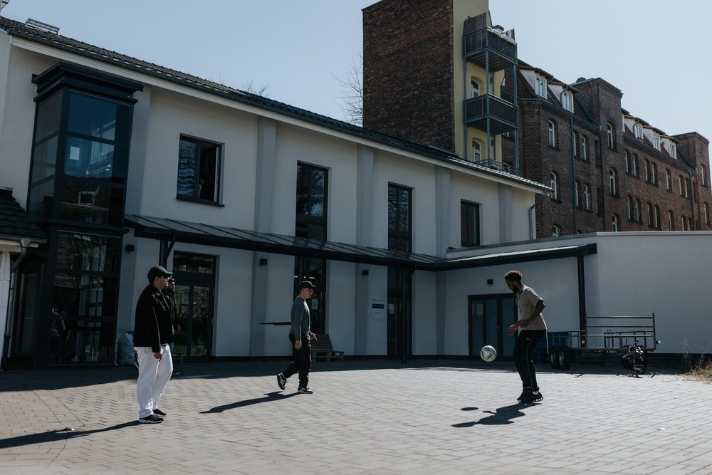 Drei Personen spielen mit einem Fußball in einem sonnigen Innenhof vor einem modernen Gebäude mit großen Fenstern.