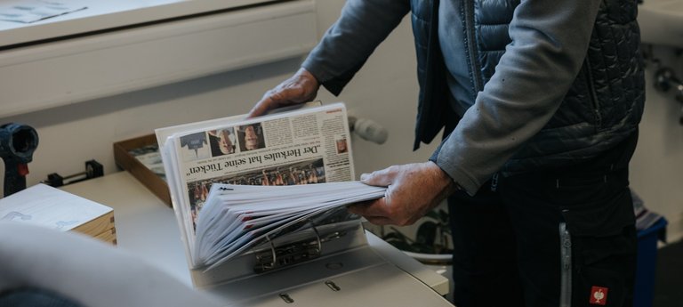 Ein Mann mit Mütze und Weste liest eine Zeitung, während er an einem Schreibtisch in einem Büro steht.
