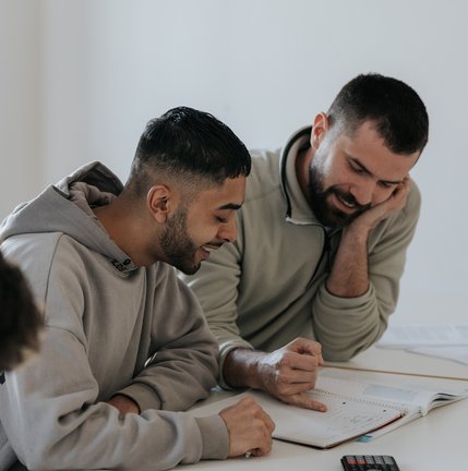 Zwei Männer, die lächelnd an einem Tisch sitzen und gemeinsam mit Notizbüchern und einem Taschenrechner lernen.