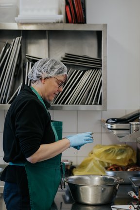 Eine Person mit Haarnetz und Handschuhen bereitet in einer Küche, neben einer Metallschüssel stehend, Essen zu.